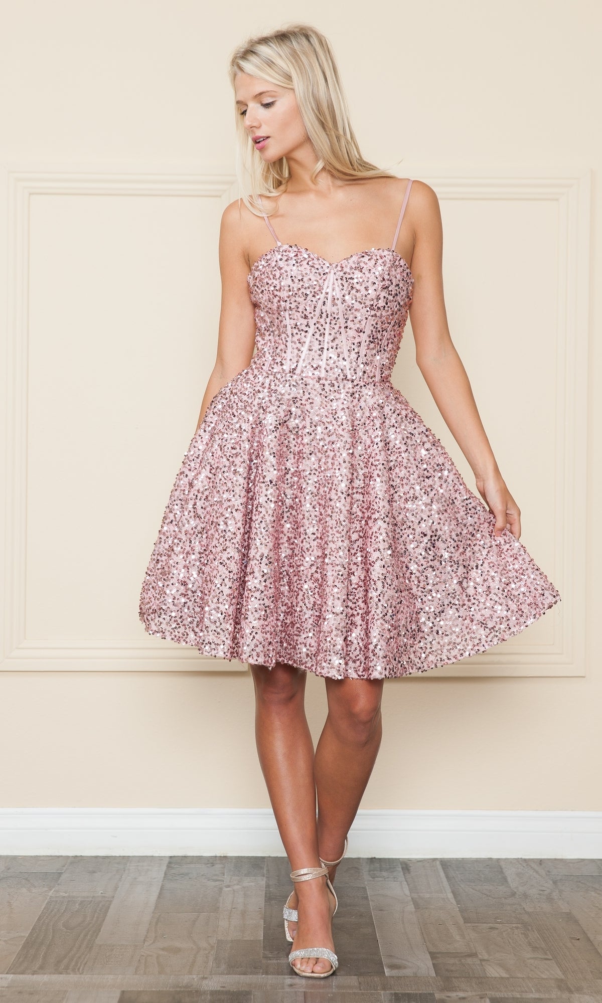 A woman stands indoors wearing the Strapless Sequin A-Line Homecoming Dress 8974 in light pink, paired with silver high-heeled sandals.